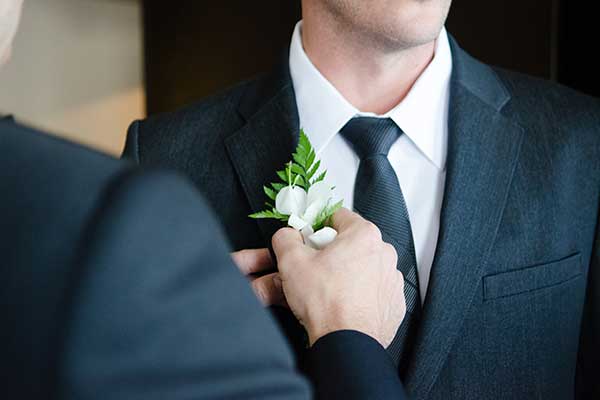 close up of man fixing a white button hole flower to the suit of another man