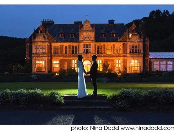 country house at night with floodlit front. Couple sand holding hands facing each other in the foreground