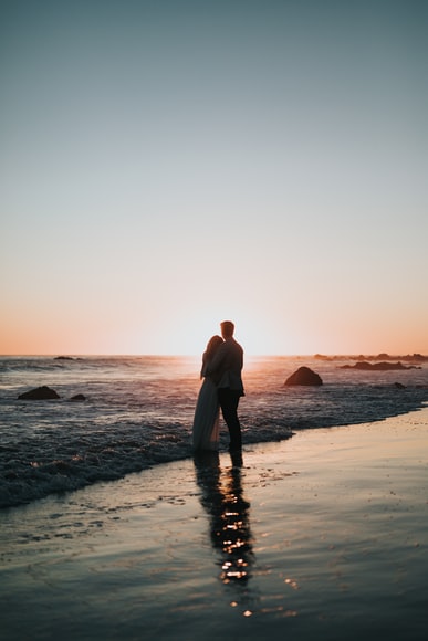 couple embracing, they are standing silhouetted in shallow breaking waves on a sandy beach with a sunset behind them.