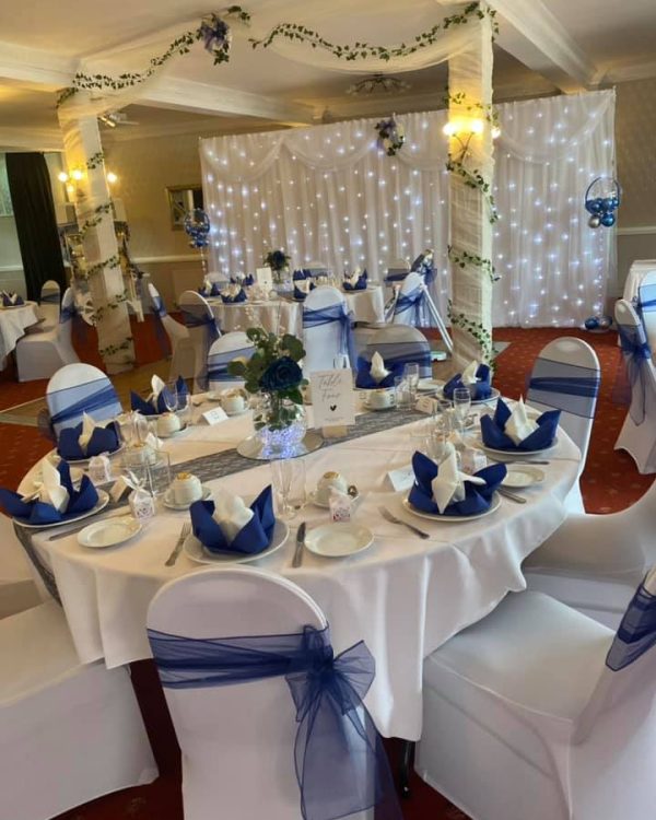 function room set up for a wedding reception. White covered chairs with dark blue sashes, round tables with coordinating blue napkins, and a flower arch with fairy lights in the background.